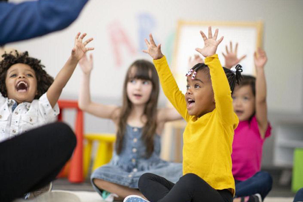 Four small children sitting on the floor of a playroom with arms raised